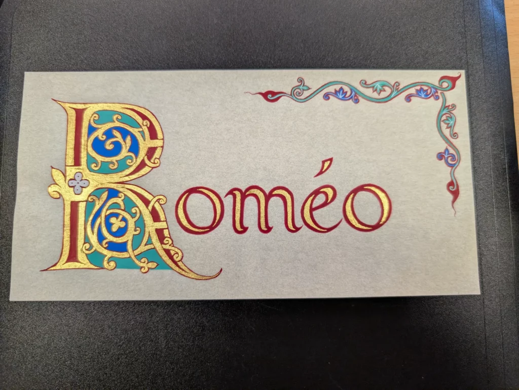 Lettrine enluminée représentant la lettre R et le reste du mot "Roméo" réalisée par un élève en cours de calligraphie.