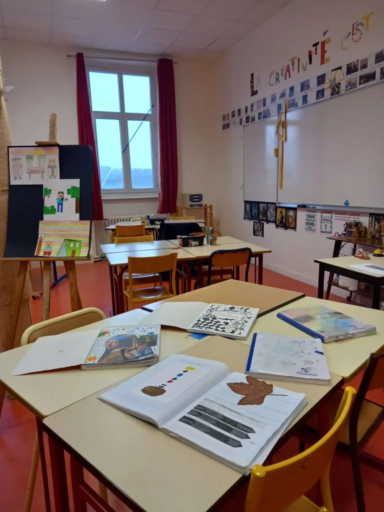 Salle d'arts plastiques lumineuse avec grandes tables de travail et productions d'élèves sur les tables.