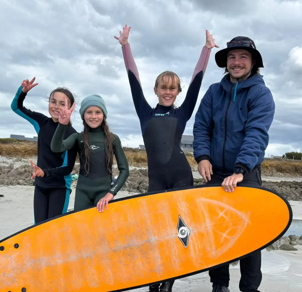 Trois élèves et un adulte tenant une planche de surf orange, en plein air.